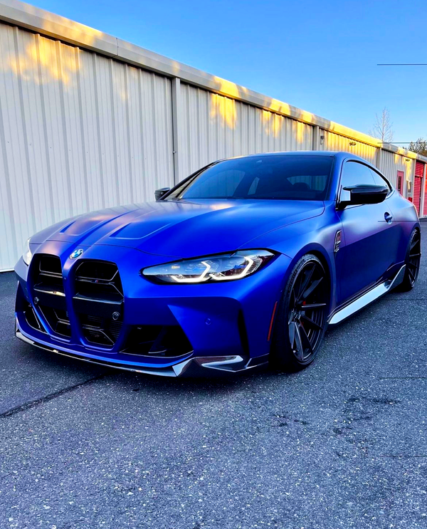 A blue sports car with sleek, angular design is parked on a paved surface. It is positioned in front of a beige industrial building under a clear blue sky.