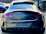 A matte black Mercedes-Benz AMG C63 parked on a paved surface, features visible exhausts and logos. Background includes parked cars and blurred greenery with a fence.