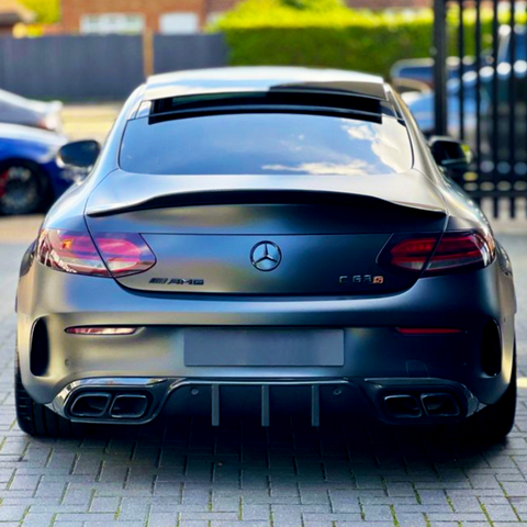 A matte black Mercedes-Benz AMG C63 parked on a paved surface, features visible exhausts and logos. Background includes parked cars and blurred greenery with a fence.