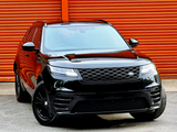 A black SUV is parked on a concrete surface, facing forward. Its sleek design reflects light, contrasting against the bright orange corrugated metal wall in the background.