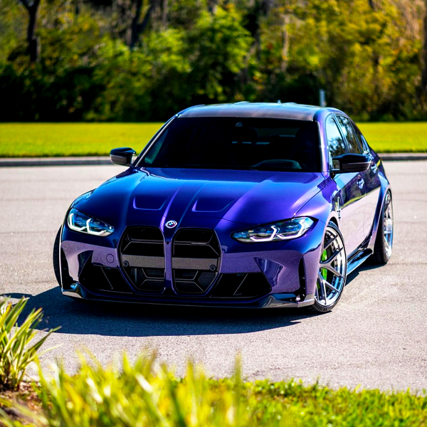 A blue sports car parked in a lot, its sleek design highlighted by sunlight, sits in front of a backdrop of grass and trees.