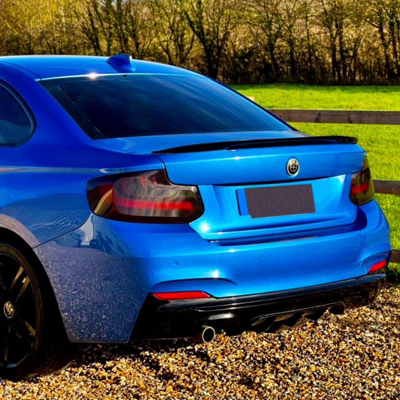 A blue car is parked on a gravel surface near a wooden fence, with a grassy field and trees in the background. The rear license plate area is blank.