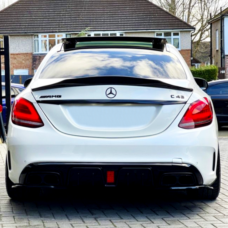 A white Mercedes-Benz AMG C43 parked on a driveway. The rear view shows badge logos and a clean, modern design. Houses and trees are visible in the background.
