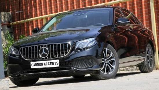 A black Mercedes-Benz car is parked on a concrete surface, reflecting a red brick wall. The grille is prominent, and the license plate reads "CARBON ACCENTS."