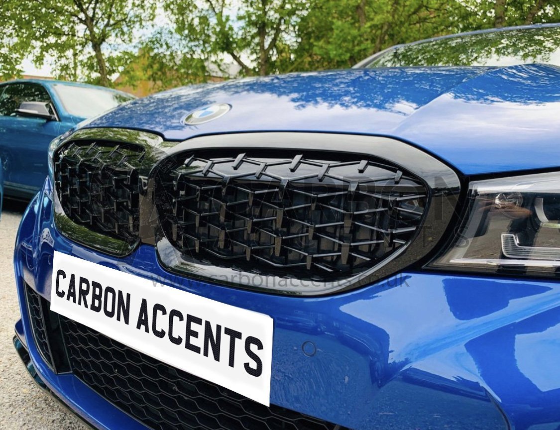 A blue car with black, angular front grille parked outdoors. Trees and another car are in the background. Text on the front reads "CARBON ACCENTS."