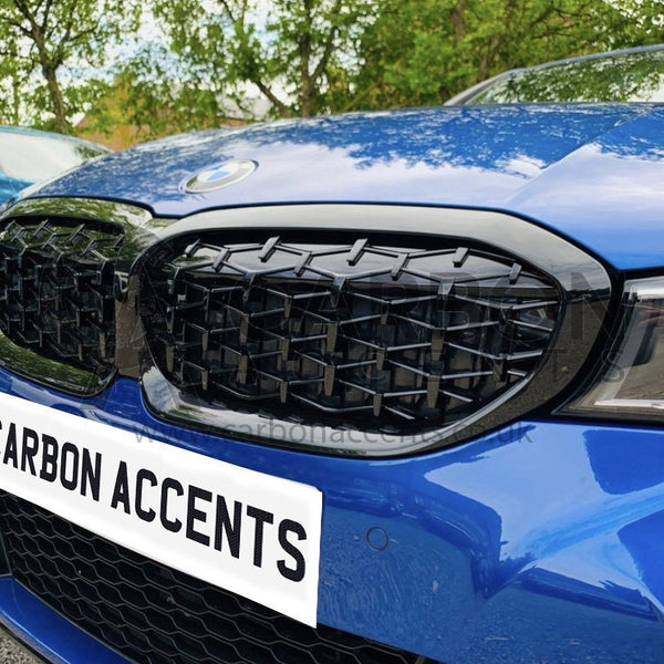 A blue car with black, angular front grille parked outdoors. Trees and another car are in the background. Text on the front reads "CARBON ACCENTS."