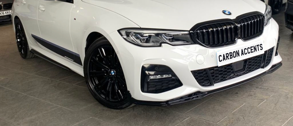 A white car with black accents is parked indoors. It features sleek, modern headlights and black wheels. The grille is prominently displayed. Text on the license plate reads "CARBON ACCENTS."