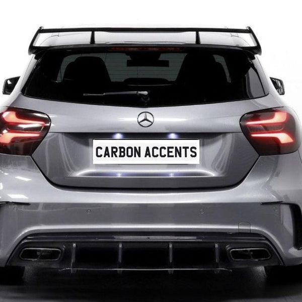 A silver car is parked in a well-lit, indoor space. It displays a license plate with the text "CARBON ACCENTS" and has a prominent rear spoiler and sleek taillights.