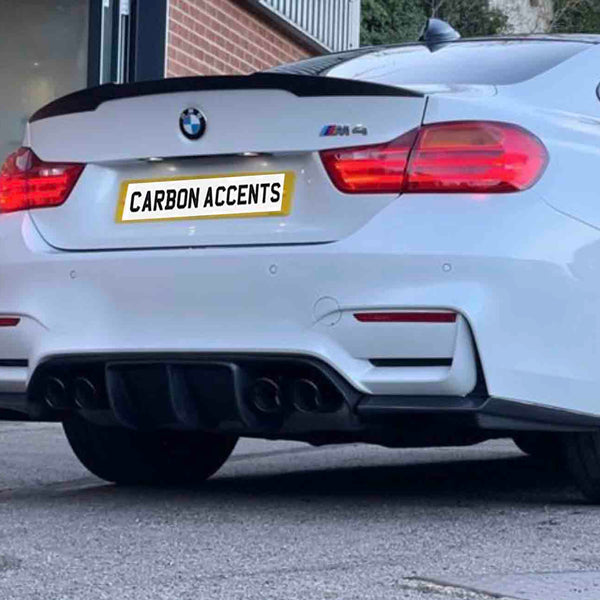 A white BMW car, parked outdoors, displays a rear spoiler and quad exhausts. The license plate reads "CARBON ACCENTS." The background includes a building entrance and greenery.