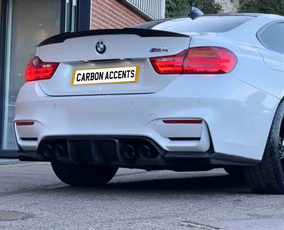 A white BMW car, parked outdoors, displays a rear spoiler and quad exhausts. The license plate reads "CARBON ACCENTS." The background includes a building entrance and greenery.
