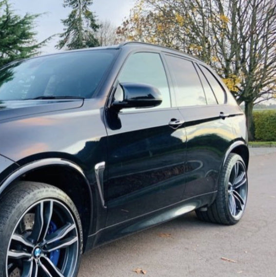 A black SUV parked on a paved surface with trees in the background, showcasing shiny, detailed wheels and reflecting the cloudy sky above.