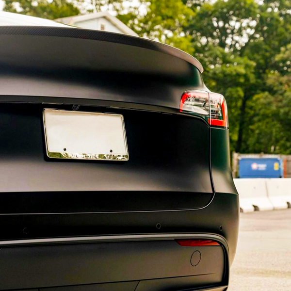A sleek black car is parked outdoors, showing its rear with a blank license plate and visible taillights. A barrier and trees are in the background.