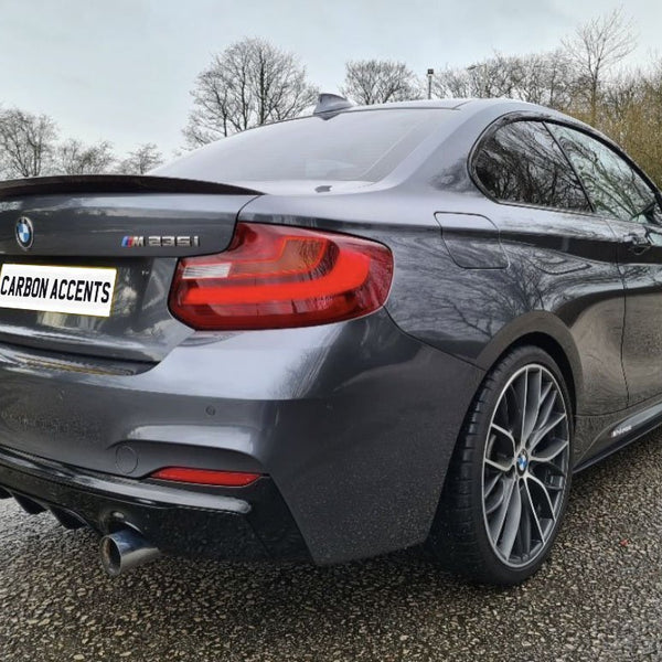 A dark gray BMW M235i is parked on a wet, textured ground with barren trees in the background. The license plate reads "CARBON ACCENTS."