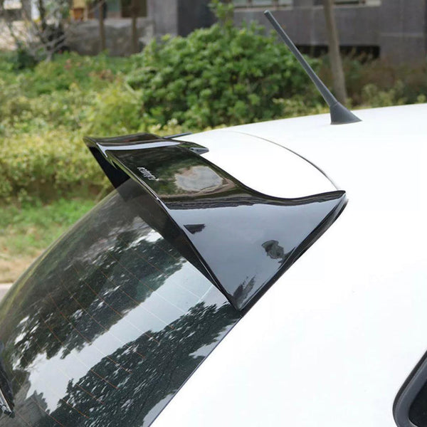 A black car spoiler is attached to the roof of a white vehicle, parked outdoors near green bushes and trees, with the word "Cuerpo" printed on the spoiler.
