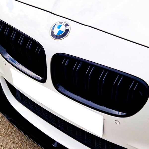 A white BMW car front displays its emblem and black kidney grille. The vehicle is stationary on a paved surface, with a blank license plate below.