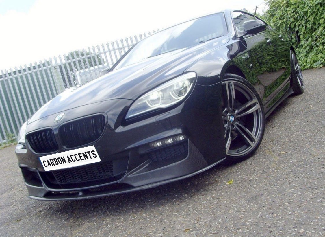 A sleek black car with custom alloy wheels parked on a paved surface, near a metal fence and greenery. The license plate reads "CARBON ACCENTS."