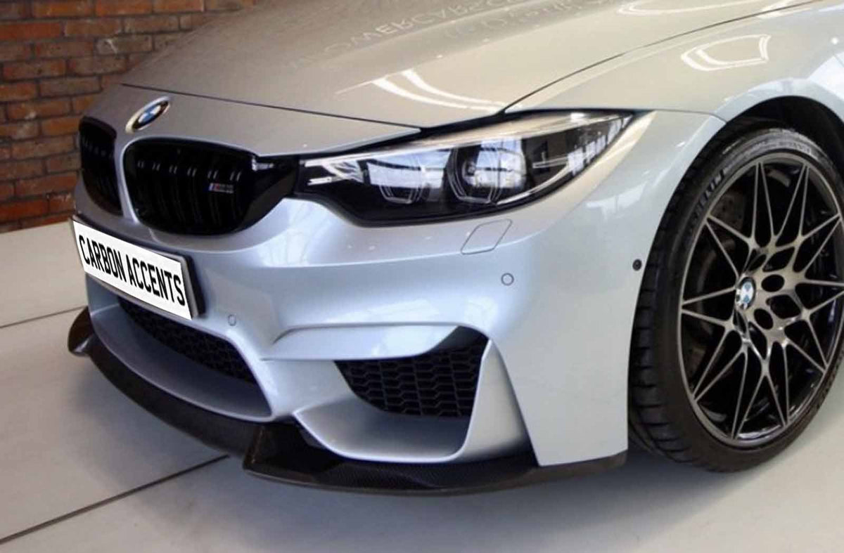A silver car front, with sleek headlights and black grille, displays a "CARBON ACCENTS" plate. It's positioned indoors against a brick wall backdrop.