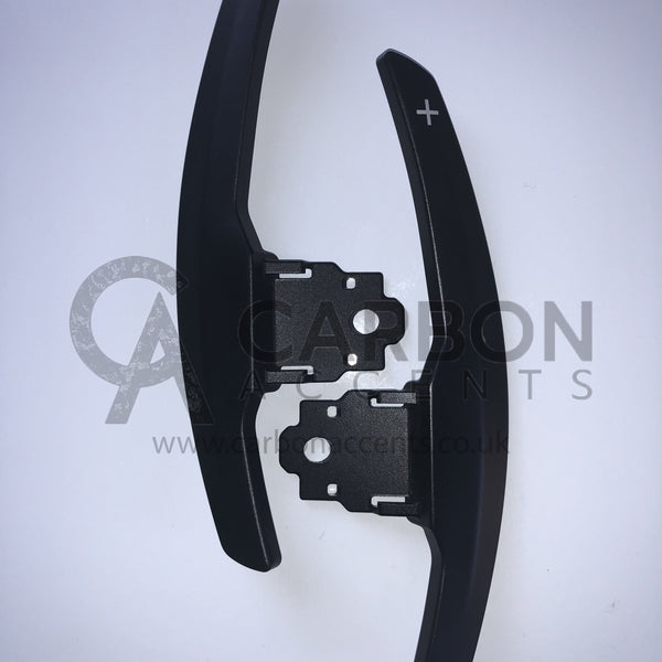 Two black paddle shifters with "+" and "-" symbols lie on a white surface. The background features text: "CARBON ACCENTS www.carbonaccents.co.uk."