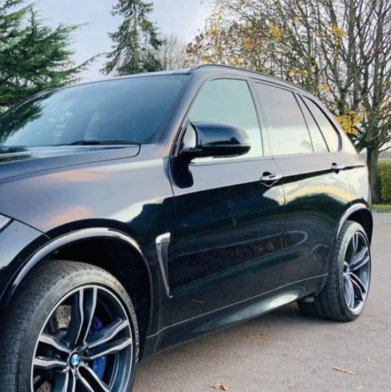 A black SUV is parked on a road. It is shiny with black alloy wheels. The background includes trees with sparse autumn leaves.