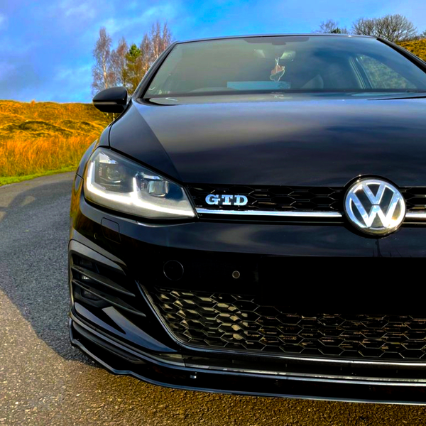 A black Volkswagen car with a "GTD" badge is parked on an asphalt road, surrounded by golden grass and trees under a blue sky.