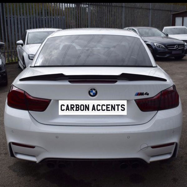A white BMW M4 is parked in a lot, displaying text "CARBON ACCENTS" on its license plate, surrounded by other parked cars and a metal fence.