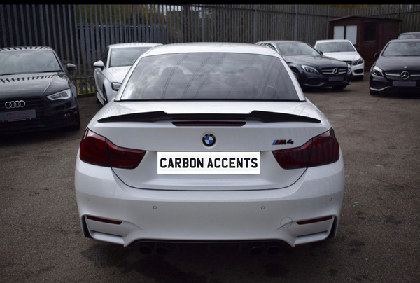 A white BMW M4 is parked in a lot, displaying text "CARBON ACCENTS" on its license plate, surrounded by other parked cars and a metal fence.