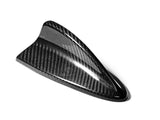 A black carbon fiber shark fin-shaped antenna is resting on a plain white background, highlighting its sleek, glossy texture and aerodynamic design.