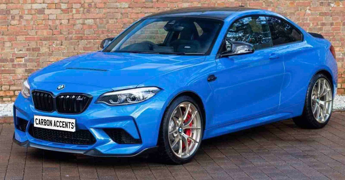 Bright blue sports car parked on brick pavement, with silver alloy wheels and tinted windows, positioned in front of a brick wall. License plate reads "CARBON ACCENTS."