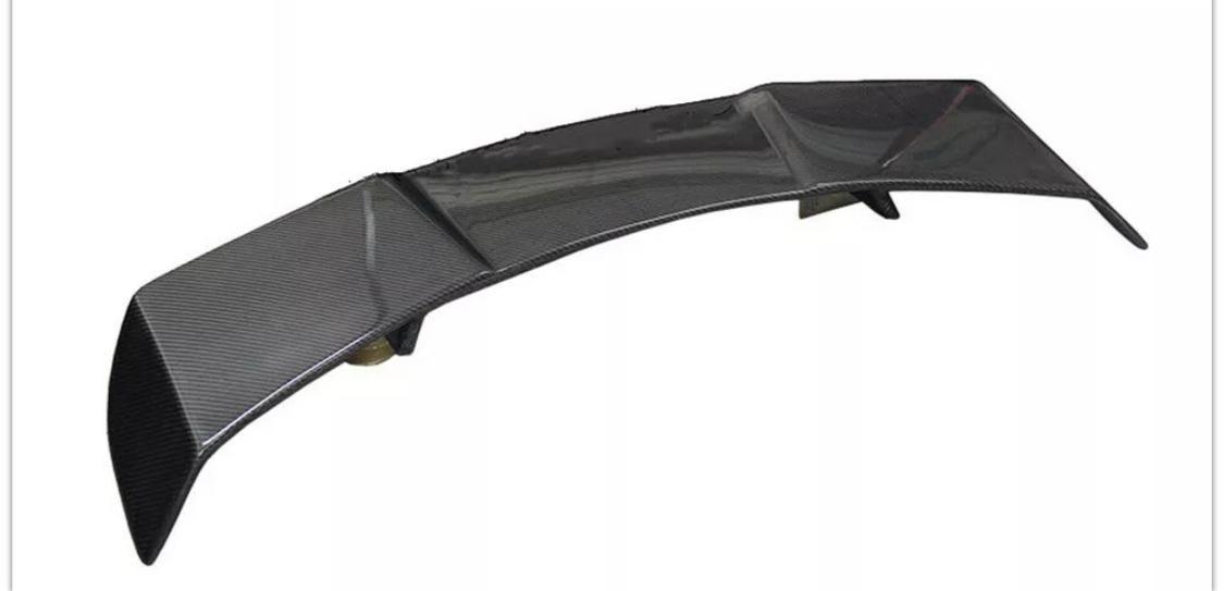 A curved carbon fiber car spoiler rests on a flat surface. The piece features aerodynamic design elements, such as a high-gloss finish and multiple support fins.