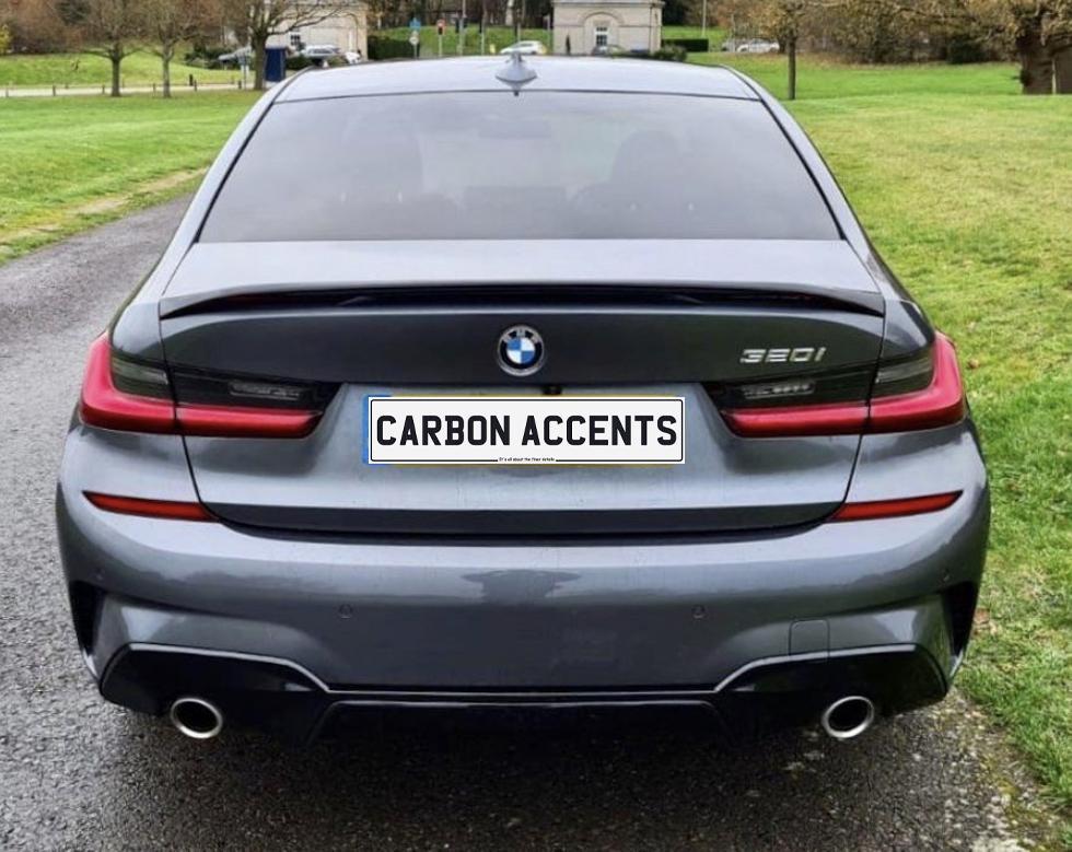 A grey BMW 3 Series car is parked on a paved road, with green grass on either side. The license plate reads "CARBON ACCENTS."