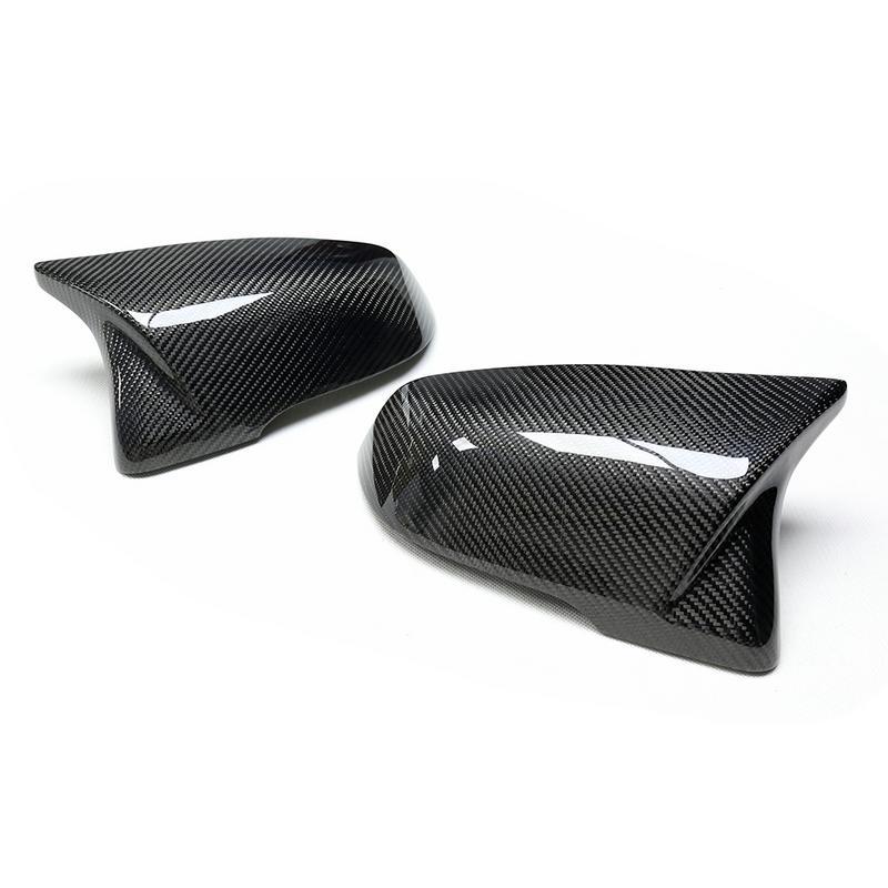 Two glossy, carbon fiber car mirror covers rest on a white surface, angled with a sleek, aerodynamic design.