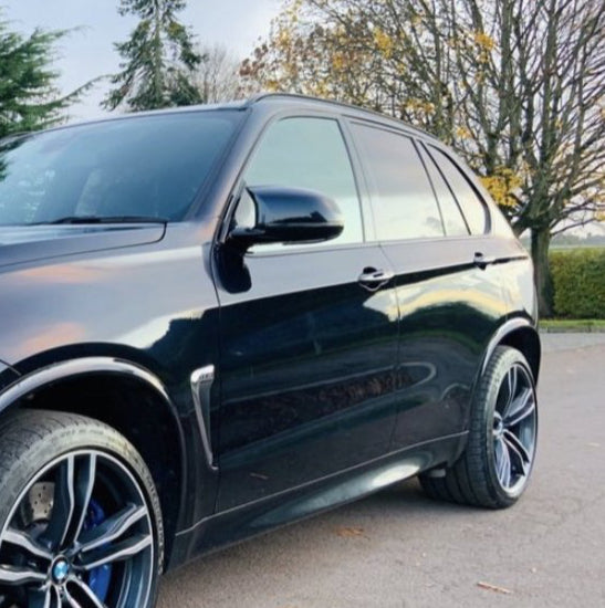 A black SUV is parked on a paved road, featuring large alloy wheels. Surrounding trees with autumn foliage and a grassy area create a serene backdrop.
