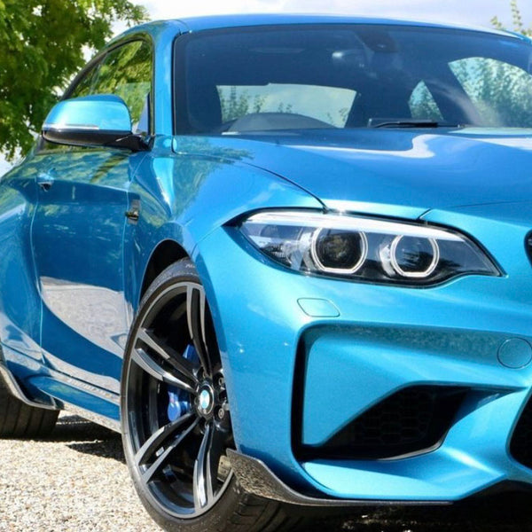 A blue sports car stands on a gravel path, with sleek rims and distinct headlights. Trees and cloudy sky form the background. License plate reads “CAR.”