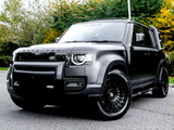 A black Land Rover Defender, parked on a paved surface, displays its modern design with bold headlights and large wheels, surrounded by trees with autumn foliage.