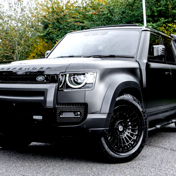A black Land Rover Defender, parked on a paved surface, displays its modern design with bold headlights and large wheels, surrounded by trees with autumn foliage.