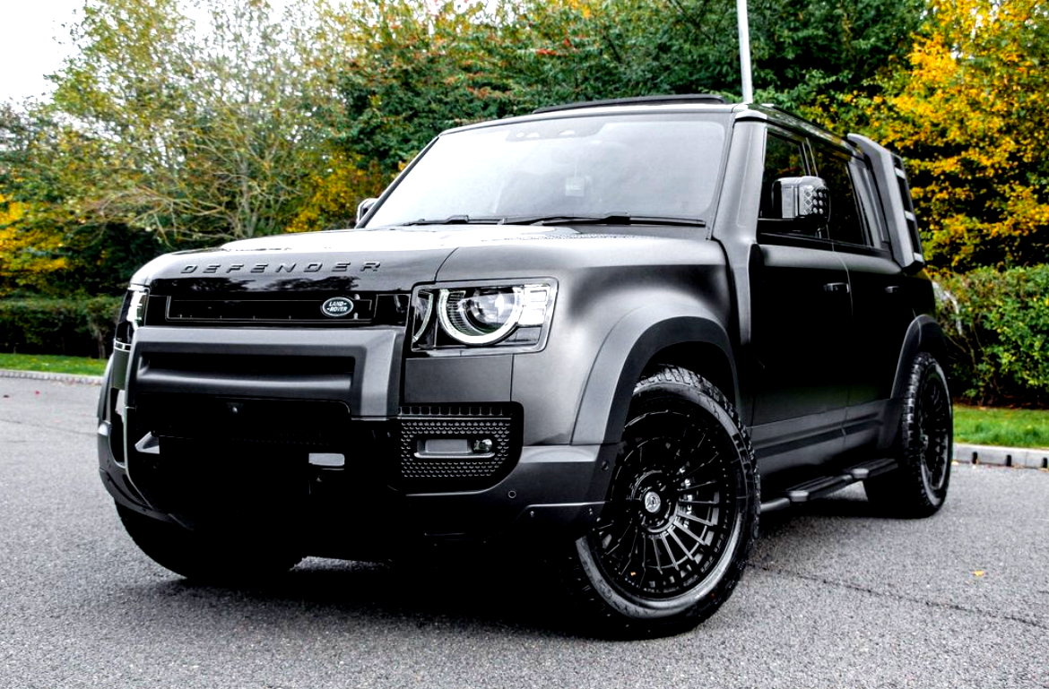 A black Land Rover Defender, parked on a paved surface, displays its modern design with bold headlights and large wheels, surrounded by trees with autumn foliage.