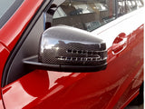A carbon fiber car's side mirror on a red vehicle reflects a residential building. Nearby windows and building details are visible, indicating an urban street environment.