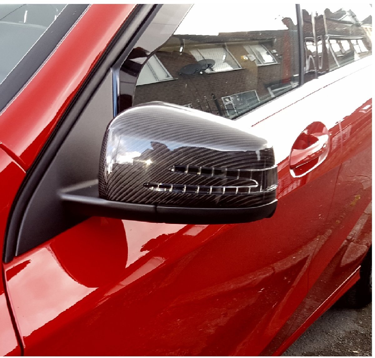 A carbon fiber car's side mirror on a red vehicle reflects a residential building. Nearby windows and building details are visible, indicating an urban street environment.