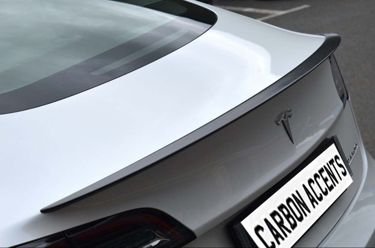 Car trunk with carbon fiber spoiler parked on asphalt, displaying logo and text "CARBON ACCENTS" on license plate. The rear design is sleek and modern.