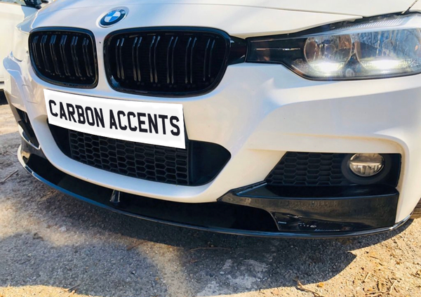 The front of a white car features a black grille and bumper accent in a sunny outdoor setting. Text reads "CARBON ACCENTS."