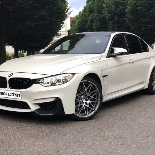 A white BMW car parked on a paved road, featuring silver alloy wheels and "CARBON ACCENTS" on the front plate, surrounded by tall, green trees and grassy areas.