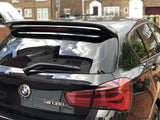 A black BMW M135i is parked, reflecting nearby houses on its shiny surface. The rear windshield wiper and spoiler are visible, with brick buildings and a driveway in the background.