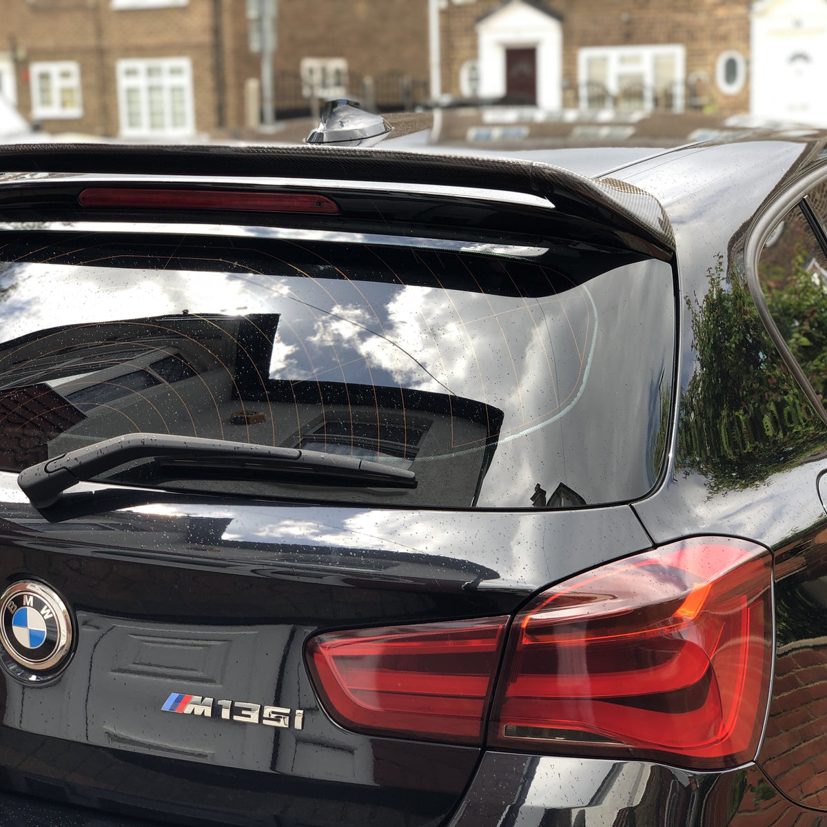 A black BMW M135i is parked, reflecting nearby houses on its shiny surface. The rear windshield wiper and spoiler are visible, with brick buildings and a driveway in the background.