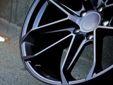 A metallic car wheel rim with a sleek, multi-spoke design rests against a textured asphalt surface, highlighting its dark, polished finish in an urban setting.