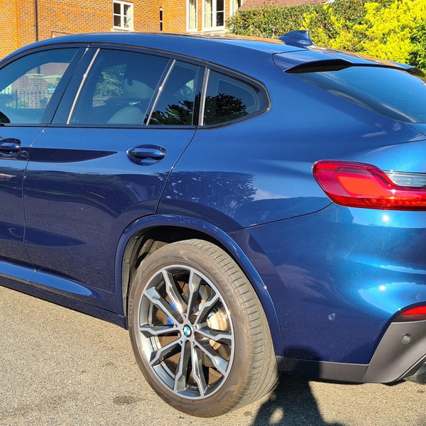 A blue BMW X4 is parked on a residential street, with sunlight highlighting its shiny surface. The license plate reads "CARBON ACCENTS." A brick house and greenery are in the background.
