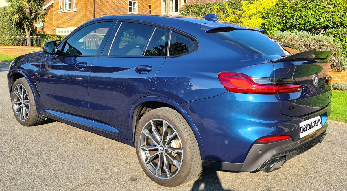 A blue BMW X4 is parked on a residential street, with sunlight highlighting its shiny surface. The license plate reads "CARBON ACCENTS." A brick house and greenery are in the background.