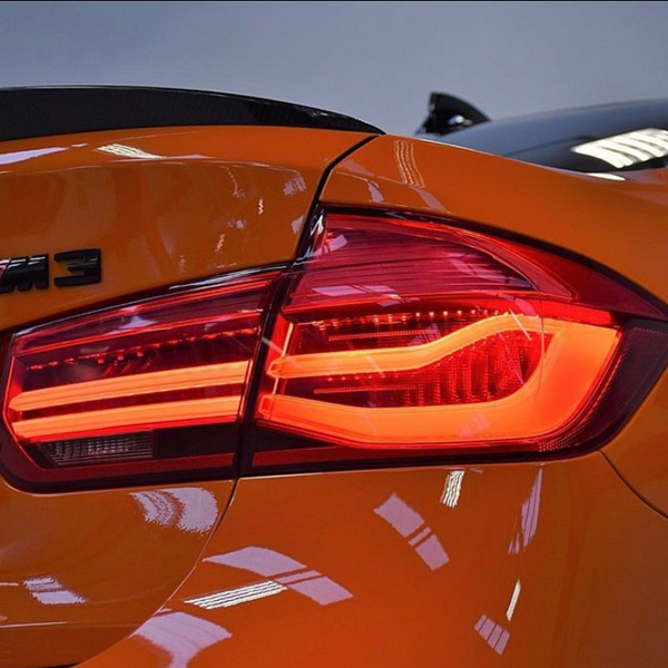 A close-up of an orange car's rear, displaying its lit brake light and an "M3" badge, parked indoors.