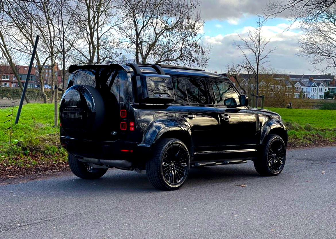 A black SUV is parked on a roadside. It's surrounded by bare trees and green grass, with houses visible in the background under a cloudy sky.