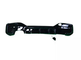A black car bumper lies flat against a white background. The bumper has several cutouts for attachments, and a small disconnected component rests nearby.