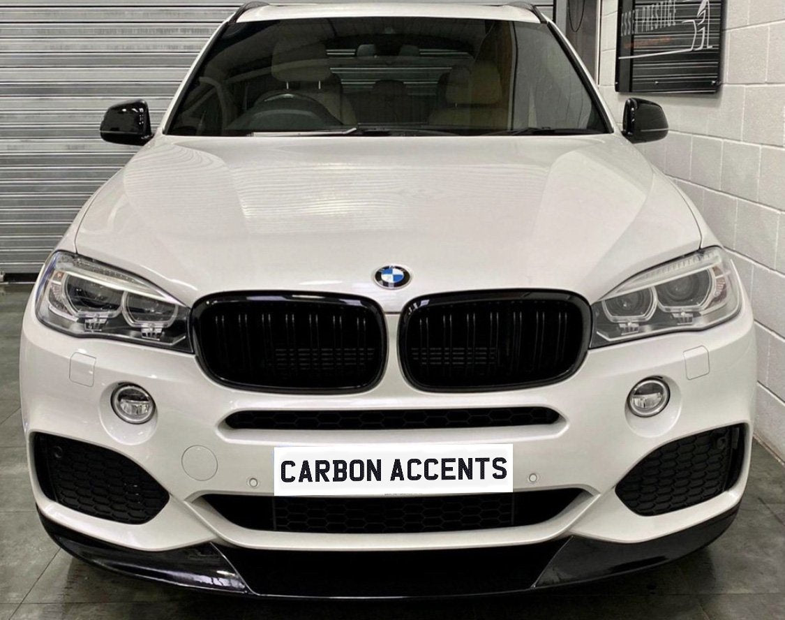 A white BMW SUV is parked indoors, facing forward with a sign on the front reading "CARBON ACCENTS." Surrounding are a tiled floor and walls with metal shuttering.
